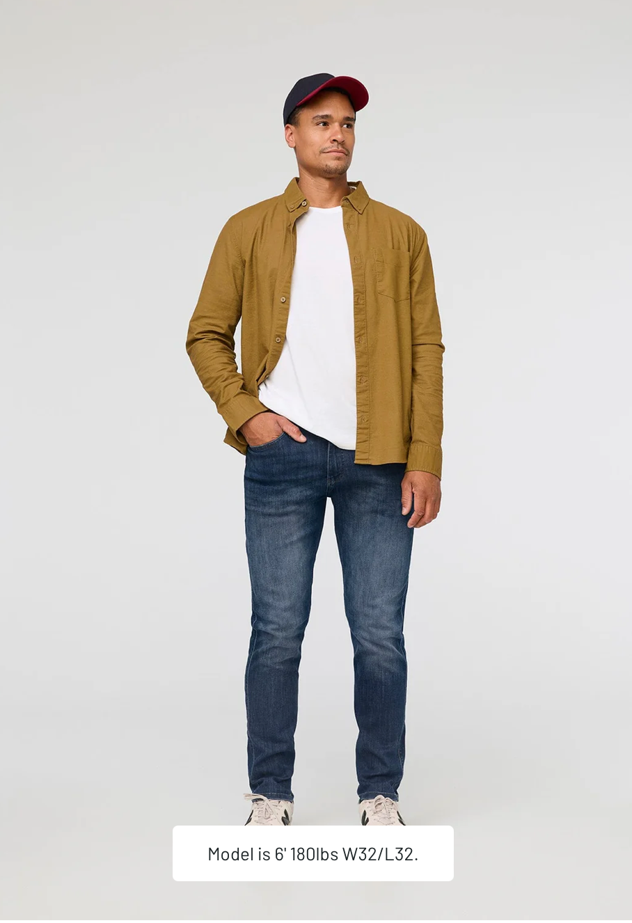 Man wearing a brown jacket, white shirt, tapered denim blue jeans, and a cap on a light gray background