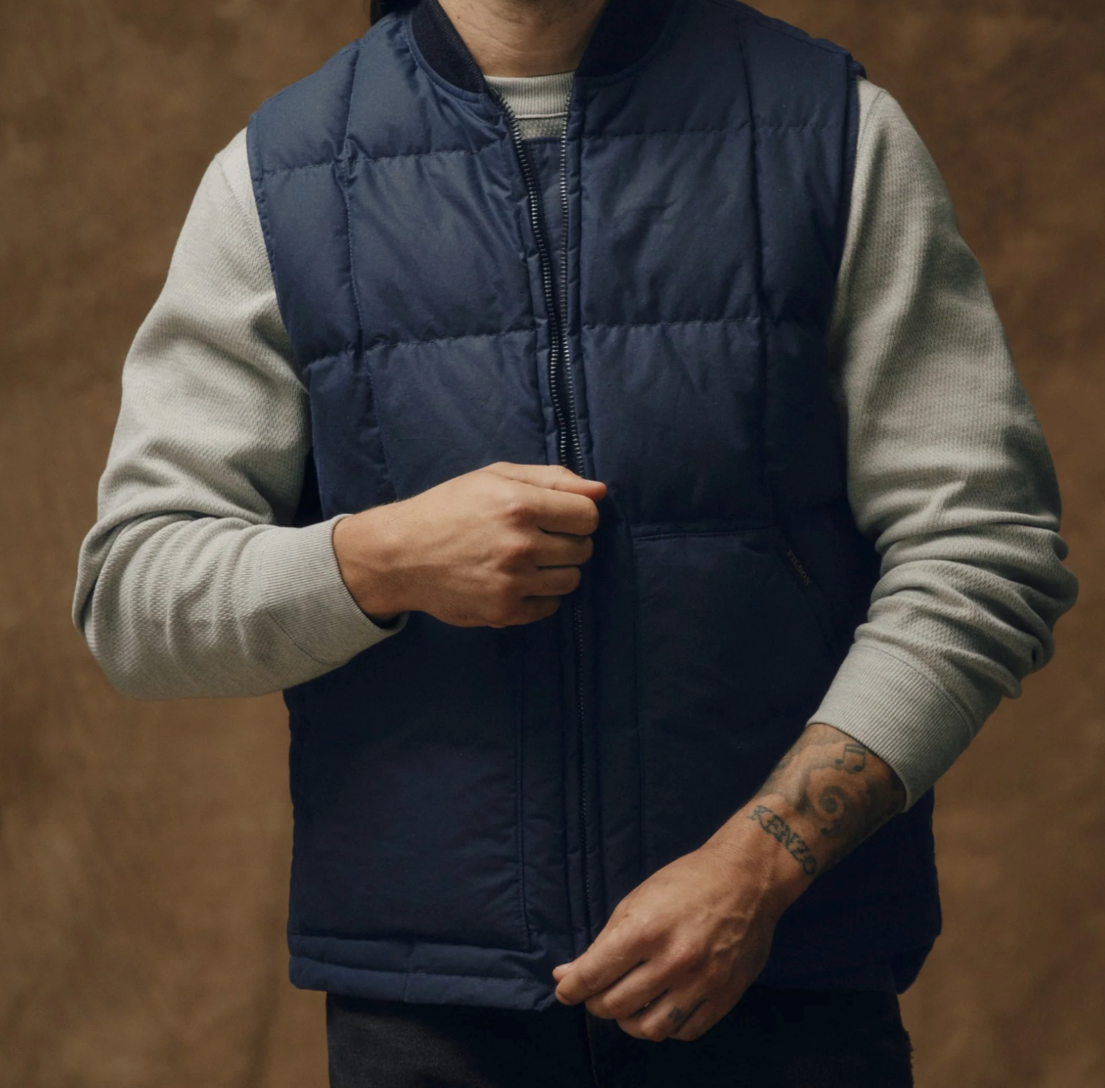 Person wearing a navy puffer vest over a light gray sweater on a brown background for men's outdoor, adventures. 