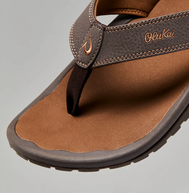 Brown sandal with 'OluKai' branding on a gray background for men's outdoor, beach days or casual wear.
