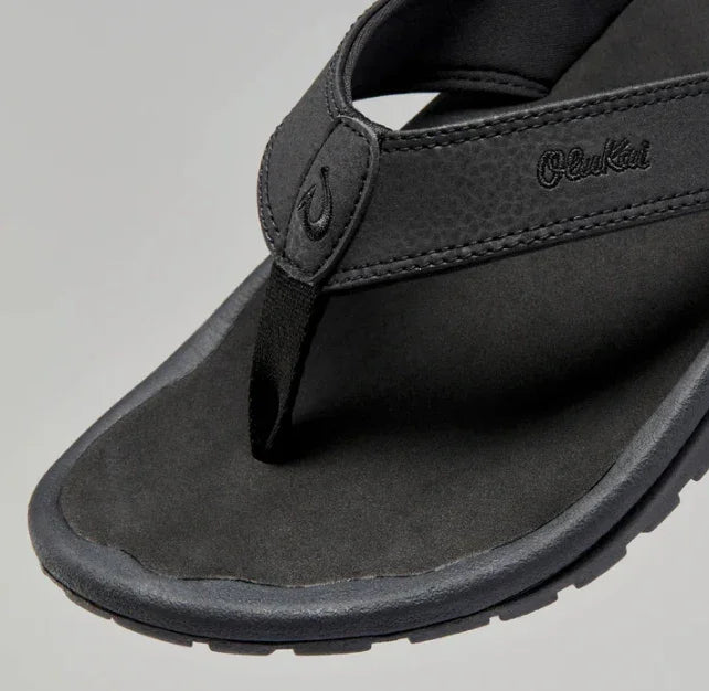 Black sandal with 'OluKai' branding on a gray background for men's outdoor, beach days or casual wear.