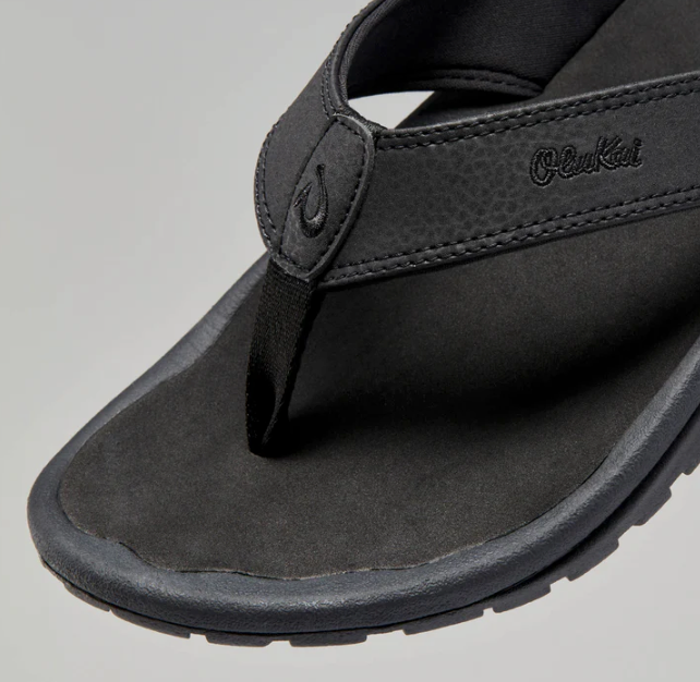 Black sandal with 'OluKai' branding on a gray background for men's outdoor, beach days or casual wear.
