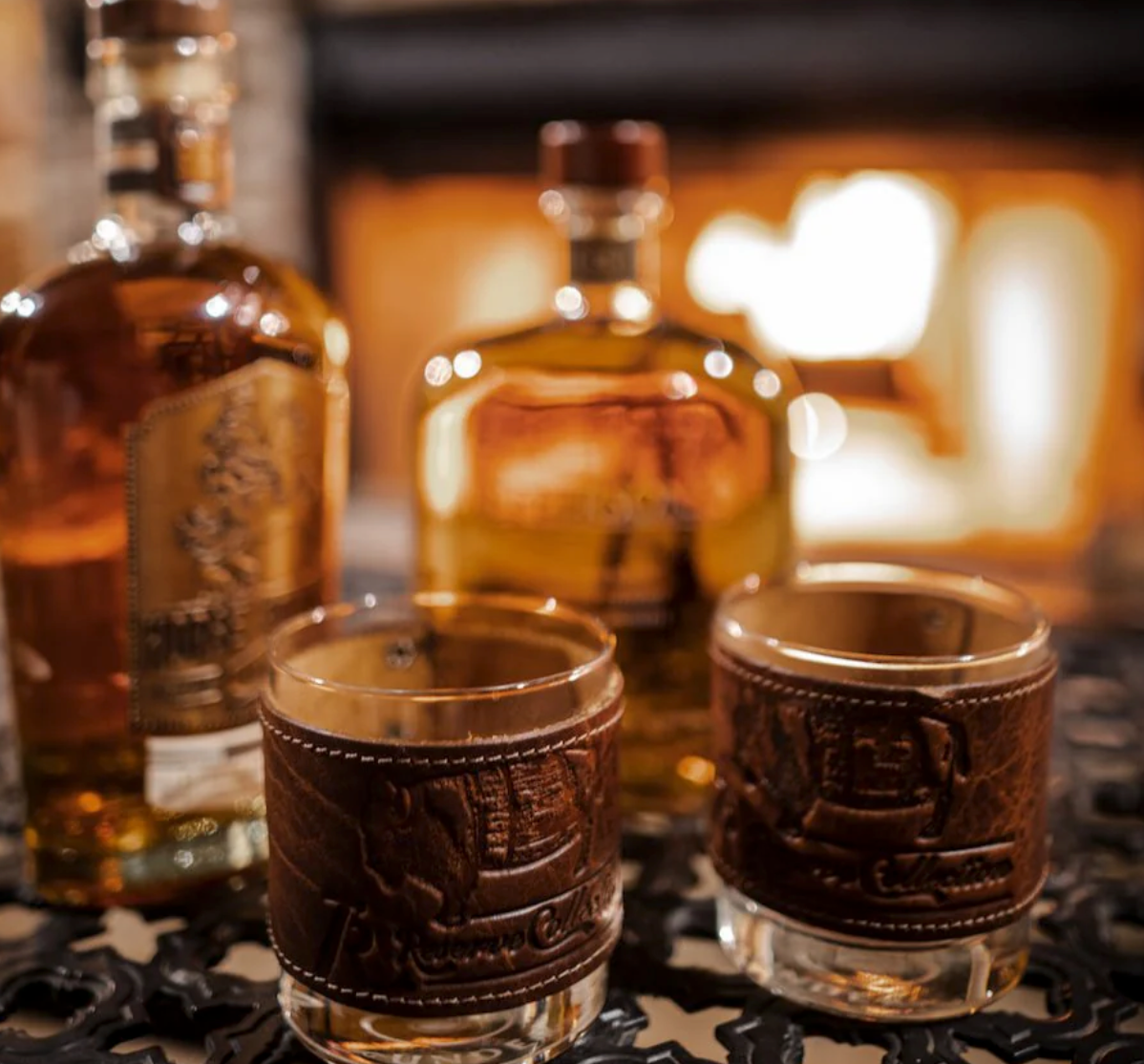 Two Over Under Bison Wrapped Bourbon Leather Grip Shot glasses with leather wraps in front of whiskey bottles on a dark surface.