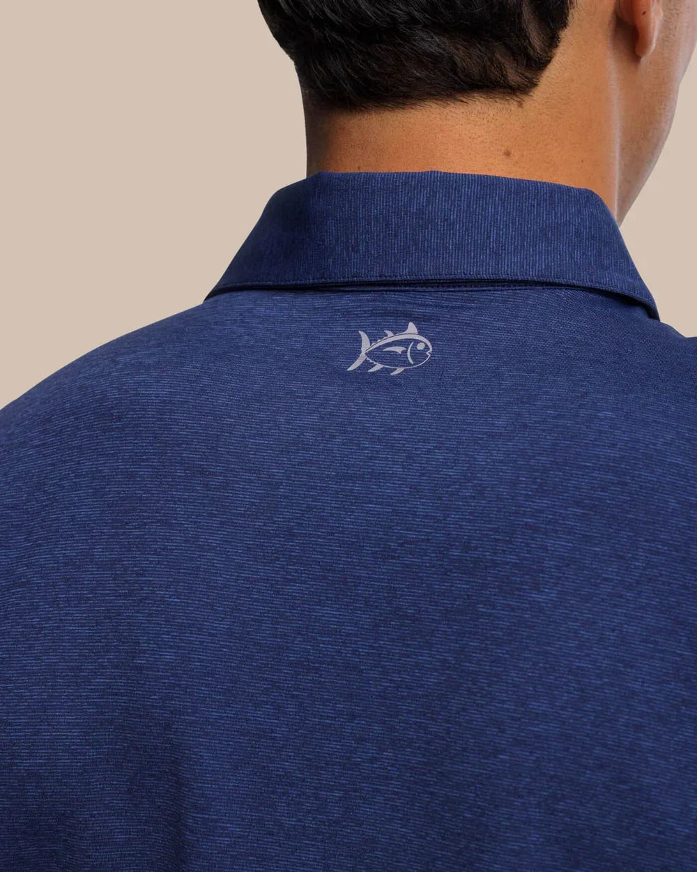 Close-up of Southern Tide Breeze Performance Heather Blue  Polo shirt with a logo on the collar against a beige background